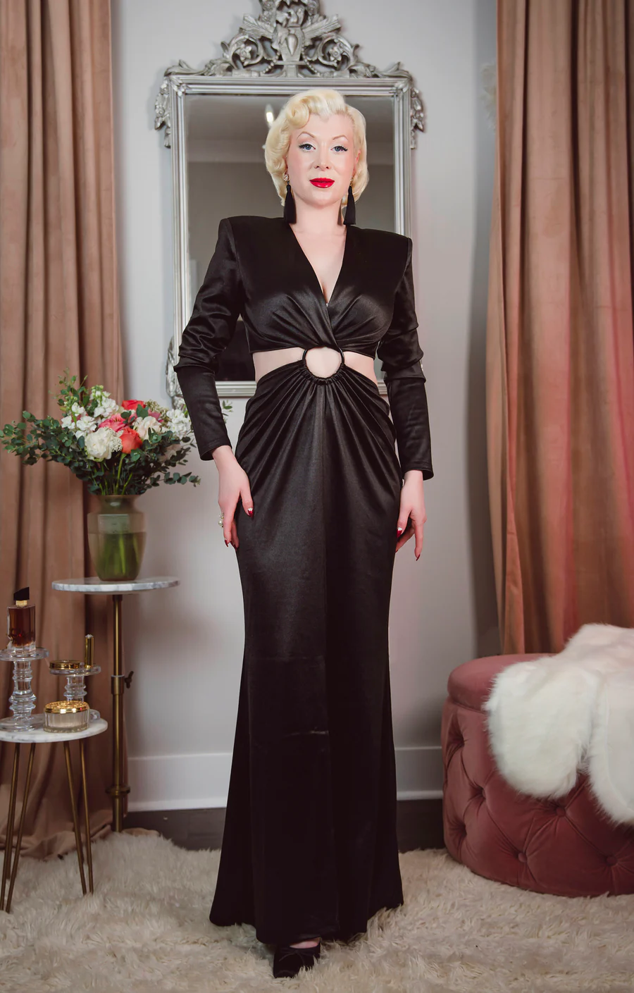 Woman in a black evening gown standing in front of a mirror in an elegant room.