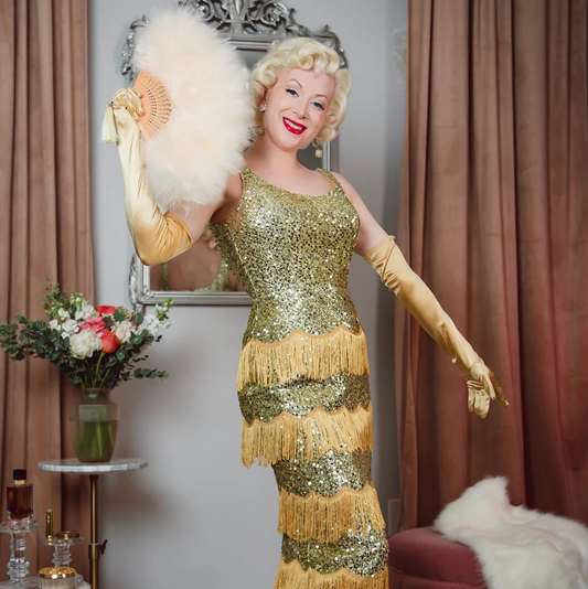 Person in a gold sequin dress with fringe holding a feathered fan, standing in a room with curtains and a mirror.