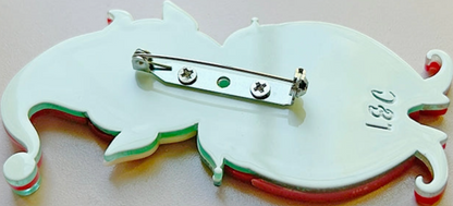 A close-up of the back of a white enamel pin shaped like a stylized animal or creature, showing a silver safety pin clasp, small green and red accents, and the engraved letters '3&1' on a light beige background.