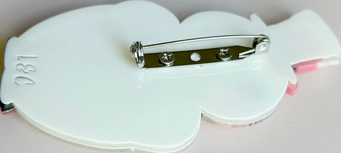 A close-up of the back of a white cloud-shaped pin-back badge or brooch, showing a silver metal safety pin clasp and a small pink backing piece, on a light neutral background.