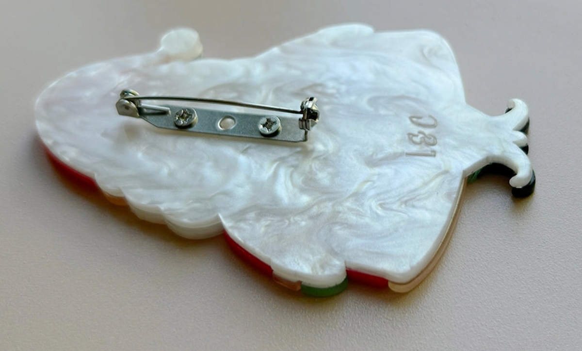 A close-up of the back of a white pear-shaped pin or brooch with a silver safety pin clasp, featuring the letters 'L&C' and small red and green accents along the edge, set against a neutral beige background.