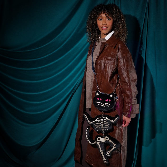 A model wearing a brown faux-leather trench coat and a beige knit sweater stands against a draped teal velvet background, holding a black cat-shaped crossbody bag with a skeleton design.