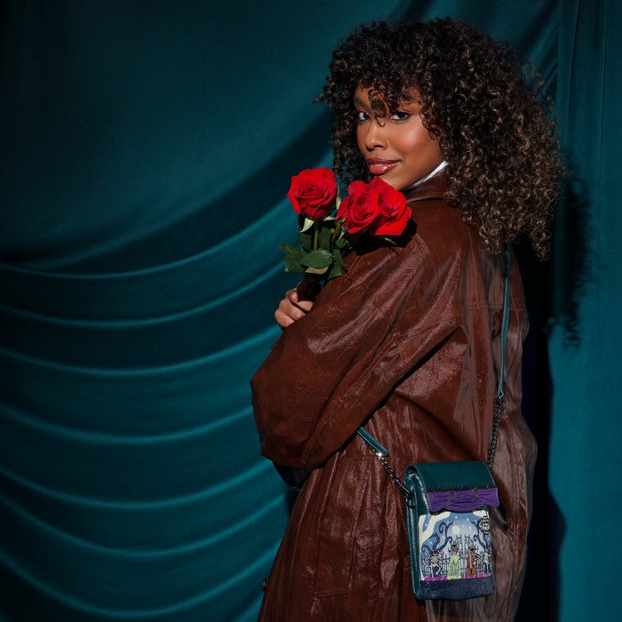 A woman with curly dark hair stands against a teal draped backdrop, wearing a brown leather jacket and holding two red roses. She carries a small dark teal crossbody bag with a colorful illustrated front pocket and a chain strap.