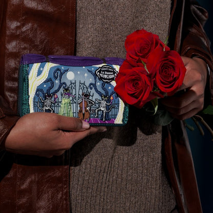 A person wearing a brown leather jacket and brown knit sweater holds a colorful illustrated zip wallet in one hand and a bouquet of three red roses in the other. The wallet features a dark fantasy illustration with purple, blue, green, and black tones.