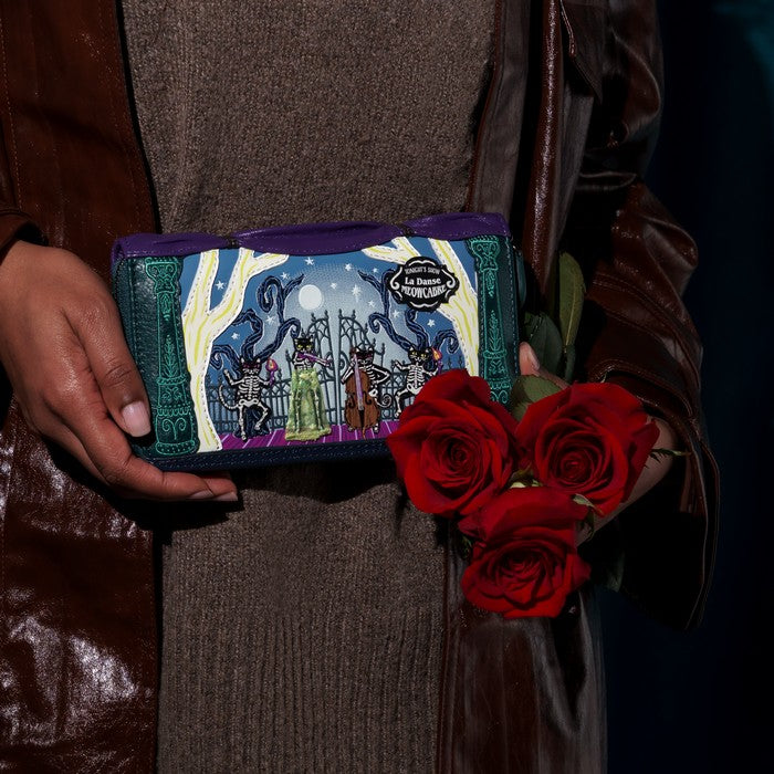 A person in a brown leather jacket holds a purple and teal illustrated zip-around wallet featuring a gothic graveyard scene, with three red roses in the other hand against a dark background.