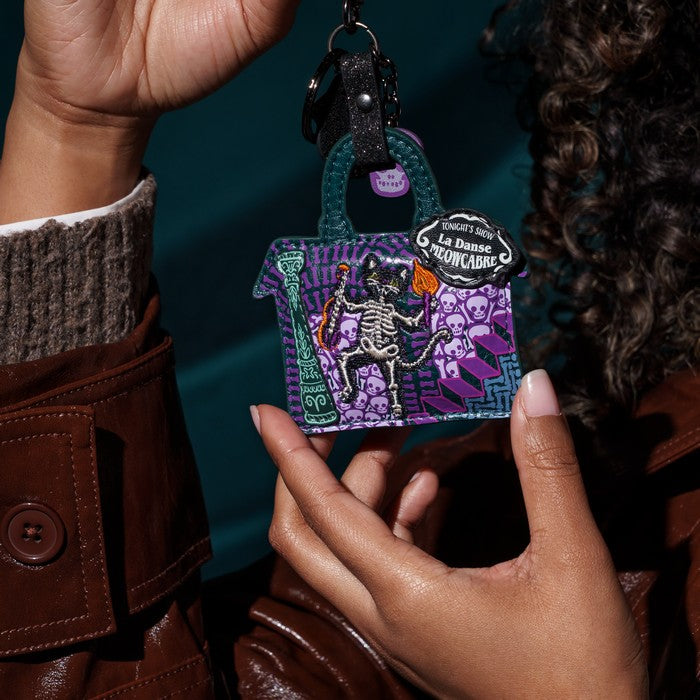 A person holds a small handbag-shaped keychain featuring a purple and black cat skeleton illustration with a lantern and a black tag that reads 'La Catrina's Mooncake.' The photo is close-up and styled with a dark teal background and brown leather clothing.