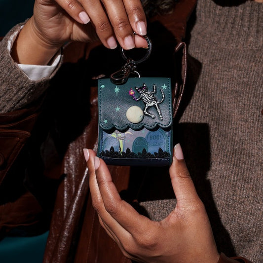A person holds a small square keychain or card holder featuring a dark teal night scene with a skeleton cat, stars, a moon, and a tombstone. The item has a black metal key ring and is shown against a brown leather jacket and brown knit sweater background.