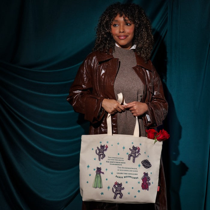 A woman stands against a dark teal draped backdrop holding a cream canvas tote bag printed with purple and green unicorn illustrations and text. She wears a shiny brown jacket over a gray turtleneck, with red roses tucked into the bag.