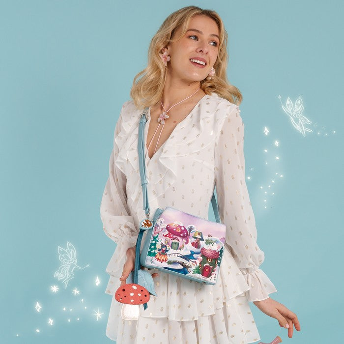 A square-shaped light blue crossbody handbag featuring a whimsical winter forest illustration with mushroom houses, a snowy bridge, and a small treehouse. The bag has a matching light blue strap and silver-tone hardware, with decorative tag accents hanging from the side.