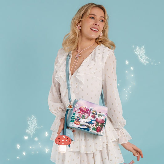 A square-shaped light blue crossbody handbag featuring a whimsical winter forest illustration with mushroom houses, a snowy bridge, and a small treehouse. The bag has a matching light blue strap and silver-tone hardware, with decorative tag accents hanging from the side.