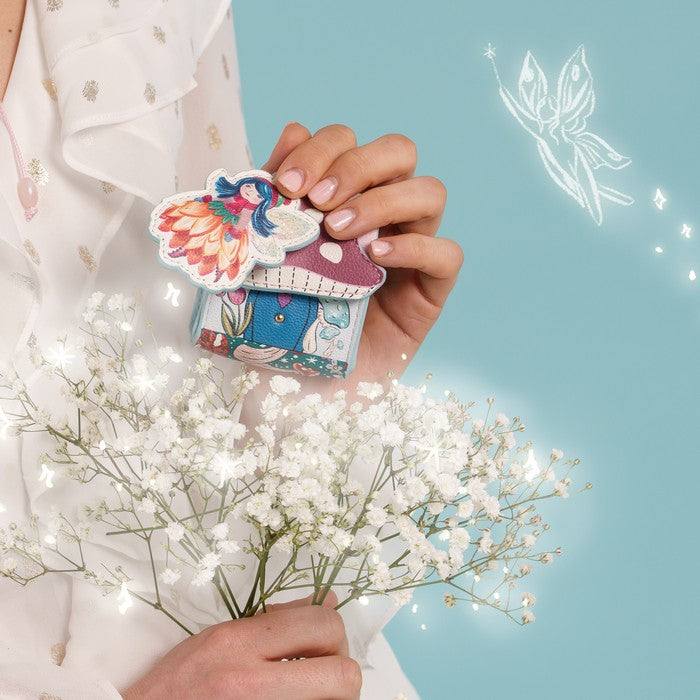 A person in a white floral dress holds a small colorful fairy-themed house charm or pin with a butterfly detail, surrounded by white baby's breath flowers against a light blue background with a glowing fairy illustration.