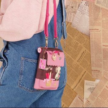 A woman wearing a pink sweater and denim overalls models a small pink crossbody bag decorated with cartoon characters, set against a background of hanging vintage newspaper pages.