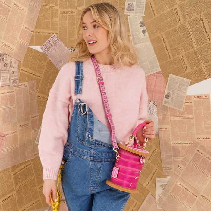 A woman wearing a pink sweater and blue denim overalls carries a bright pink cylindrical bucket bag with a pink strap, posed against a background of vintage newspaper clippings.