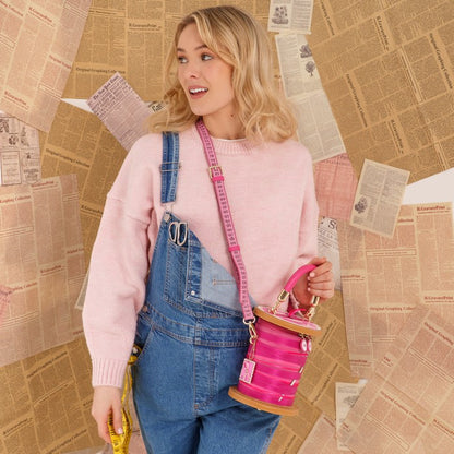 A woman wearing a pink sweater and blue denim overalls carries a bright pink cylindrical bucket bag with a pink strap, posed against a background of vintage newspaper clippings.