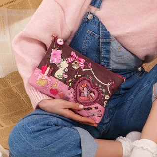 A woman sits cross-legged on newspaper-covered flooring, wearing a pink sweater and denim overalls, holding a pink Hello Kitty patterned pouch. Yellow measuring tape, scissors, and a coffee cup are scattered around her.