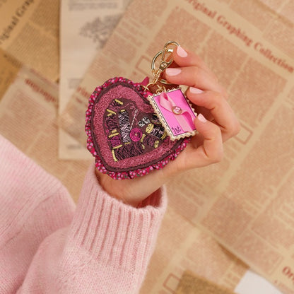 A hand holding a heart-shaped keychain purse with a miniature pink suitcase charm, set against a background of vintage newspaper pages.