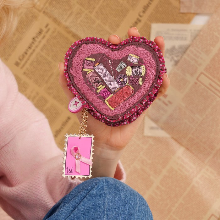A hand holds a pink heart-shaped embroidered coin purse decorated with sewing notions and a dangling pink tag, set against a background of newspaper pages.