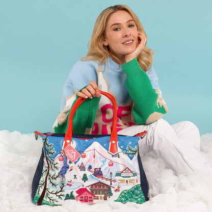 A woman sits on white faux fur against a light blue studio background, holding a large tote bag with a colorful winter mountain village print and red handles.