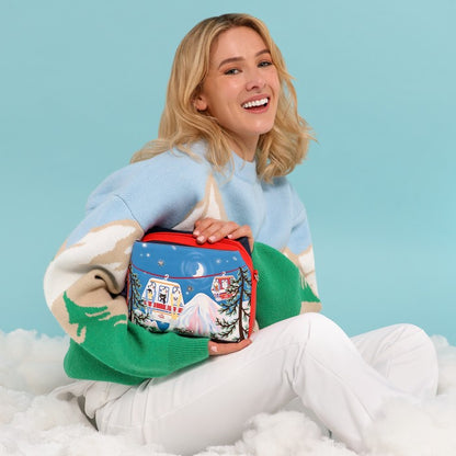 A woman sitting on white fluffy snow wears a light blue, green, and cream knit sweater while holding a small zippered pouch decorated with a winter mountain scene. The pouch has a red zipper and is set against a solid light blue background.