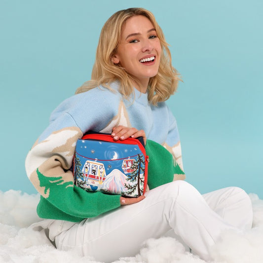 A woman sitting on white fluffy snow wears a light blue, green, and cream knit sweater while holding a small zippered pouch decorated with a winter mountain scene. The pouch has a red zipper and is set against a solid light blue background.