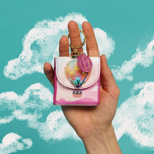 Hand holding a colorful keychain pouch with a hot air balloon design against a blue sky with clouds.