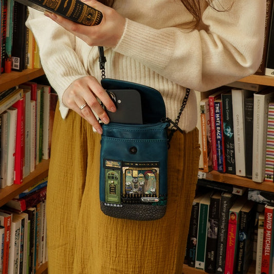Person holding a blue crossbody bag with a phone case in front of a bookshelf.