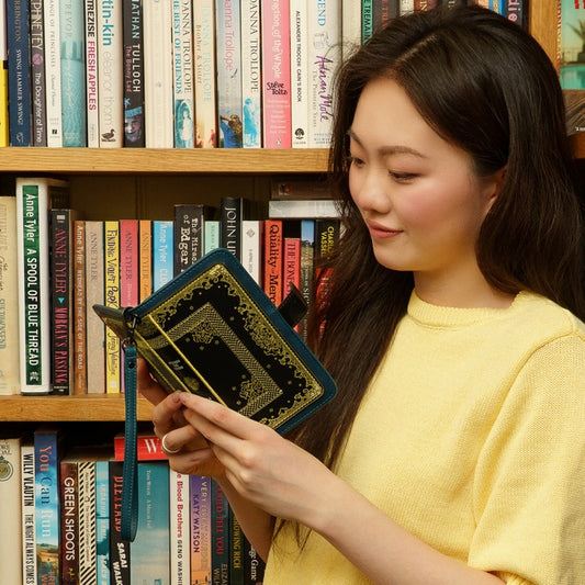 Woman holding a small book or notebook in front of a bookshelf filled with books.