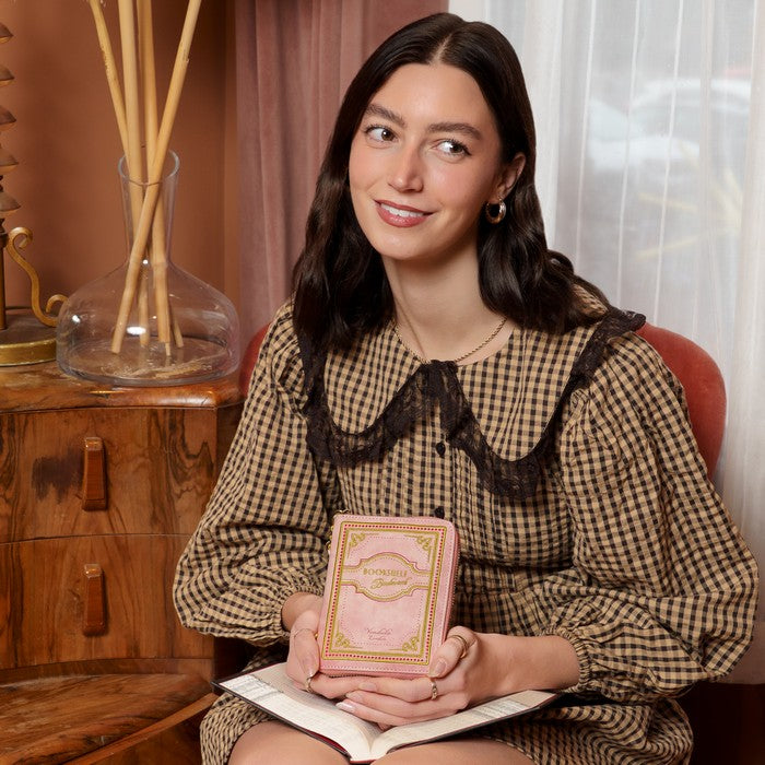 Woman holding a pink and gold box in a room with wooden furniture and curtains.
