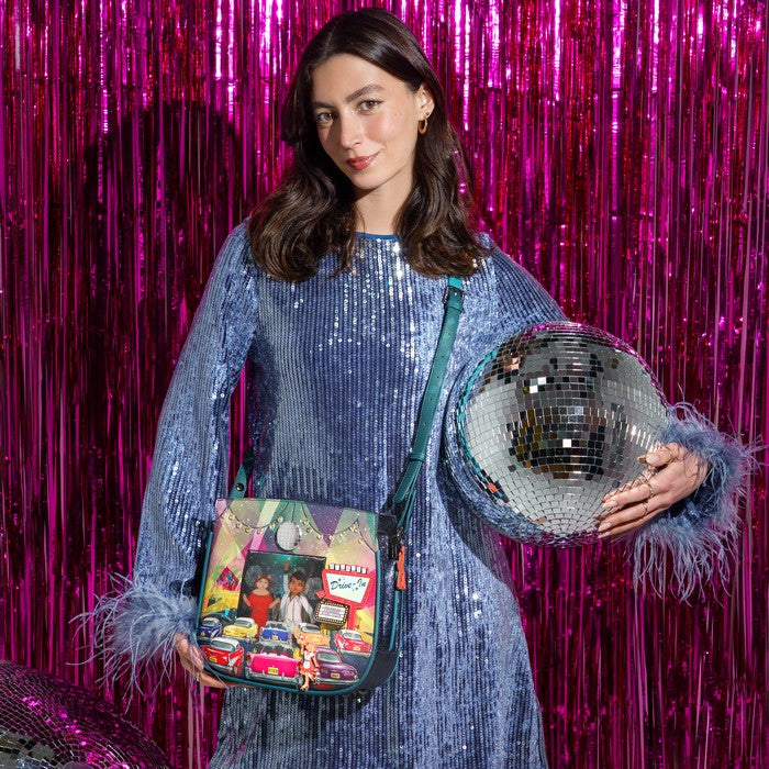 A woman in a blue sequined dress holds a colorful crossbody bag with a retro car print and a large silver disco ball, set against a bright pink tinsel curtain.