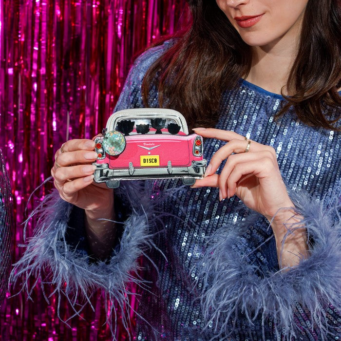 A woman in a blue sequined dress holds a pink vintage car enamel pin with a yellow 'B15CM' license plate, set against a shiny pink tinsel backdrop.