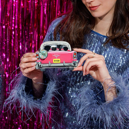 A woman in a blue sequined dress holds a pink vintage car enamel pin with a yellow 'B15CM' license plate, set against a shiny pink tinsel backdrop.