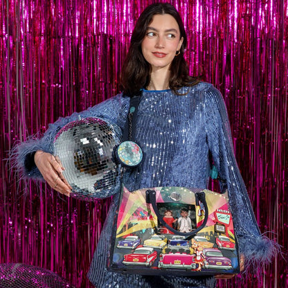 A woman in a blue sequined dress holds a colorful vintage-style tote bag with illustrated cars and a disco ball charm, set against a bright pink tinsel backdrop.