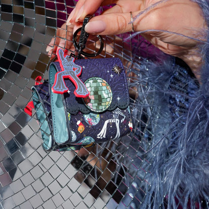 A hand holds a small black keychain bag decorated with a disco ball, stars, and a white cat illustration. The bag features a black strap and is adorned with a red and gray alien charm, set against a reflective silver disco ball mirror background.