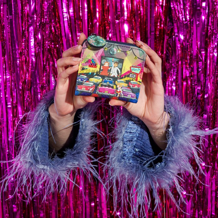 A person holds a small illustrated wallet or clutch in front of a shiny pink tinsel backdrop. The wallet has a colorful vintage storefront scene with cars and a green circular embellishment near the top.