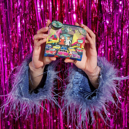 A person holds a small illustrated wallet or clutch in front of a shiny pink tinsel backdrop. The wallet has a colorful vintage storefront scene with cars and a green circular embellishment near the top.