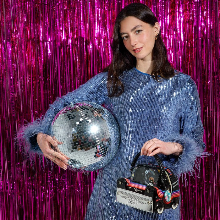 A woman stands in front of a bright pink tinsel backdrop, wearing a blue sequined dress with feathered cuffs and holding a large silver disco ball and a small black car-shaped handbag.