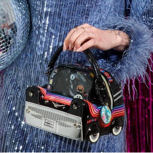 A hand holding a small novelty handbag shaped like a retro car with rainbow stripes, shown against a person wearing a blue sequined jacket and a disco ball in the background.