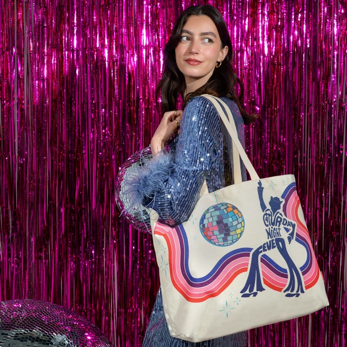 A woman wearing a sparkly blue sequined top carries a cream canvas tote bag with a colorful retro disco-themed graphic featuring a dancing figure, a disco ball, and the text 'Dancing Night Ever'. She is posed in front of a bright pink metallic tinsel curtain backdrop.