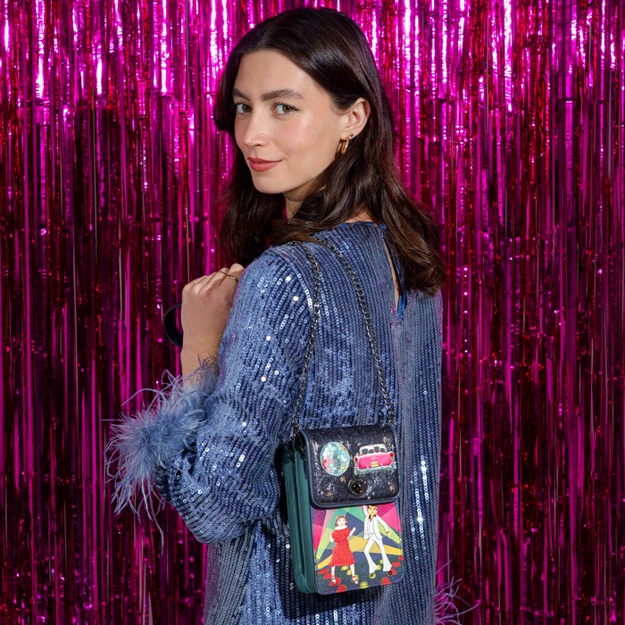 A woman wearing a blue sequined dress with feather details carries a small black crossbody bag decorated with colorful patches and a green leather trim, set against a bright pink tinsel backdrop.