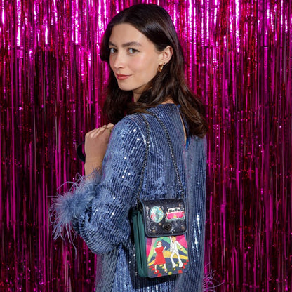 A woman wearing a blue sequined dress with feather details carries a small black crossbody bag decorated with colorful patches and a green leather trim, set against a bright pink tinsel backdrop.