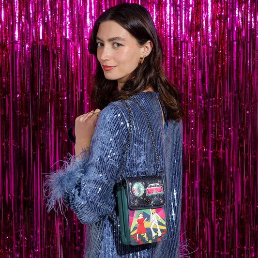 A woman wearing a blue sequined dress with feather details carries a small black crossbody bag decorated with colorful patches and a green leather trim, set against a bright pink tinsel backdrop.