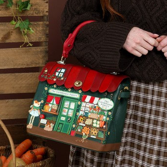 A smiling young woman with long red hair stands against a brown studio backdrop wearing a dark brown cable-knit sweater and a brown-and-white plaid skirt. She is holding a small handbag designed to look like a cozy village shop with a red roof, green facade, and colorful illustrated storefront details.