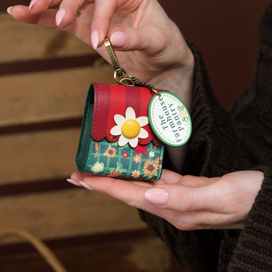 A close-up of a person holding a small floral wallet or keychain pouch. The product has a red and green striped top flap, a green floral patterned body, a white daisy flower with a yellow center, and a gold key ring with a round green tag reading 'The Farmhouse Parfums'.