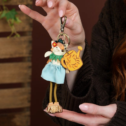 A close-up of a person holding a whimsical keychain featuring a fox character in a light blue dress holding a yellow sun-shaped object, set against a dark brown sweater and wooden background.