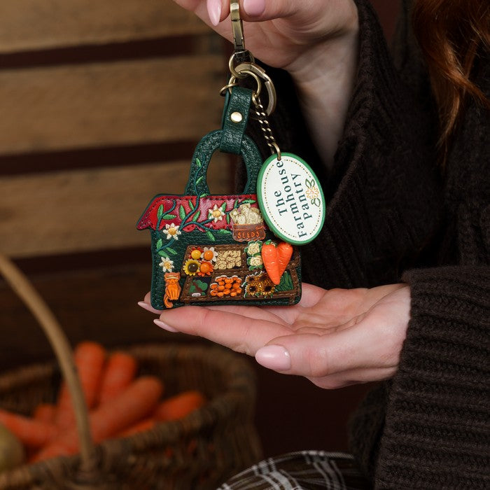 A person holds a small decorative keychain shaped like a market produce stand. The keychain has a dark green base with colorful raised details of vegetables, flowers, and fruits, plus a round white tag that reads 'Aloha from the Garden.