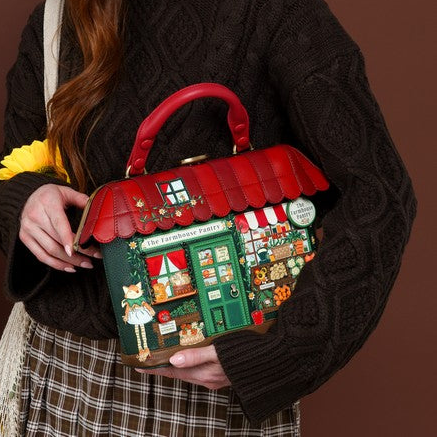A woman with long red hair holds a decorative Christmas-themed handbag shaped like a shop front. She is wearing a dark cable-knit sweater and a plaid skirt, standing against a warm brown studio background with a wooden crate and potted plants to the side.