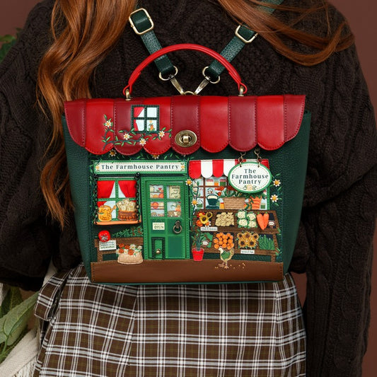A person wearing a dark brown knit sweater and a brown plaid skirt carries a decorative handbag shaped like a colorful storefront. The bag has a red top handle and scalloped red upper panel, dark green sides, and an illustrated shop front with green door, red-and-white awning, flowers, and produce displays.