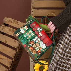 A woman with long red hair stands in a studio setting wearing a dark brown cable-knit sweater and a brown-and-beige plaid skirt. She is holding a green Christmas advent bag decorated with red gifts and green ornaments, positioned next to rustic wooden crates and sunflowers against a solid brown backdrop.