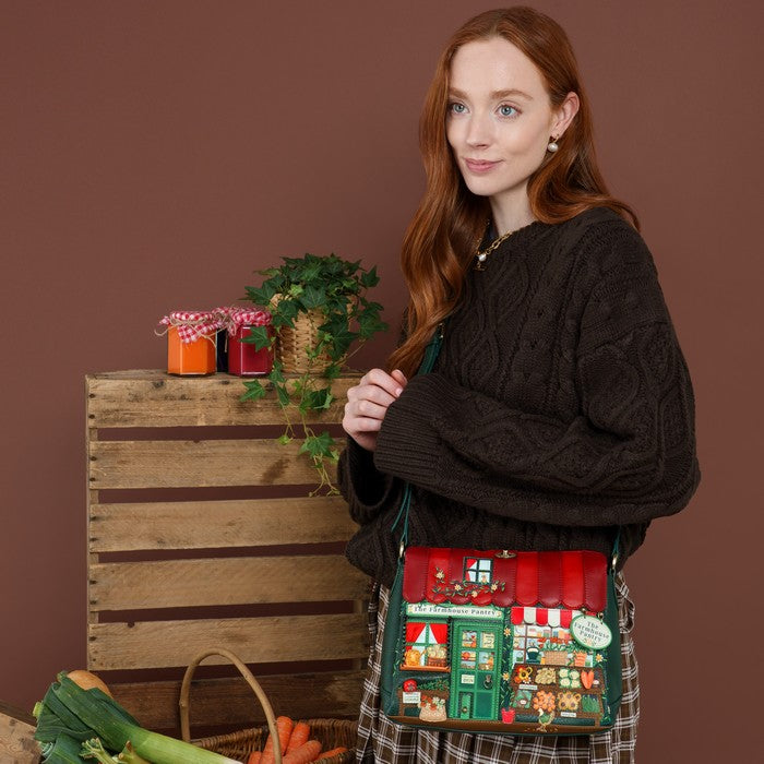A woman with long red hair models a Christmas-themed crossbody bag featuring a village shop design, set against a rustic brown background with a wooden crate and vegetables.