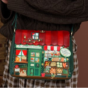 A woman with long red hair models a Christmas-themed crossbody bag featuring a village shop design, set against a rustic brown background with a wooden crate and vegetables.
